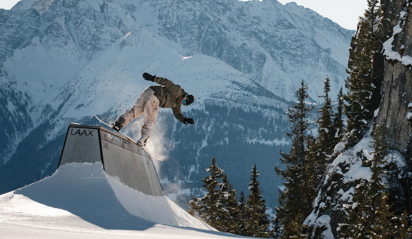 Snowboarder effectue une figure sur une box, en arrière-plan le massif de signina.