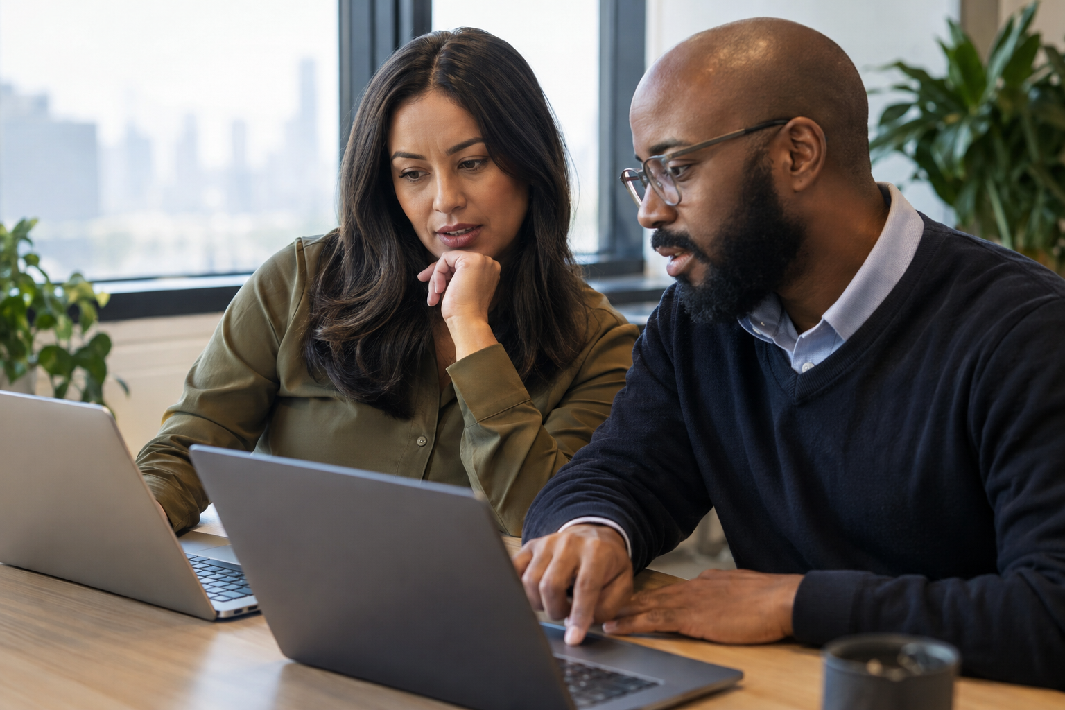 A procurement lead and IT administrator compare digital approval tools using laptops at a shared workstation.