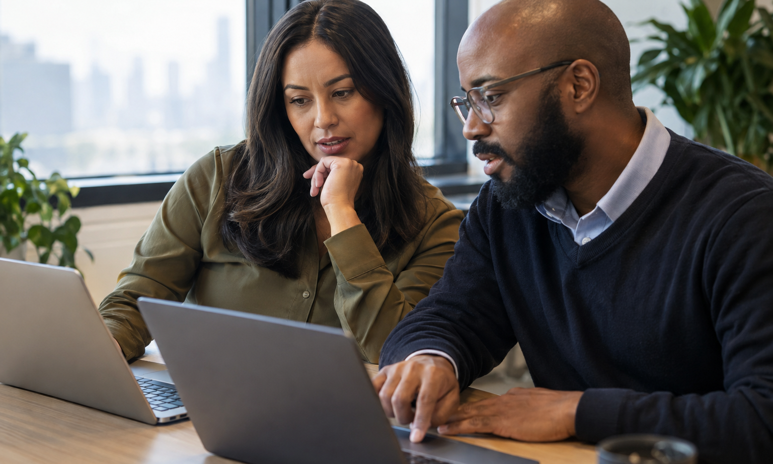 A procurement lead and IT administrator compare digital approval tools using laptops at a shared workstation.