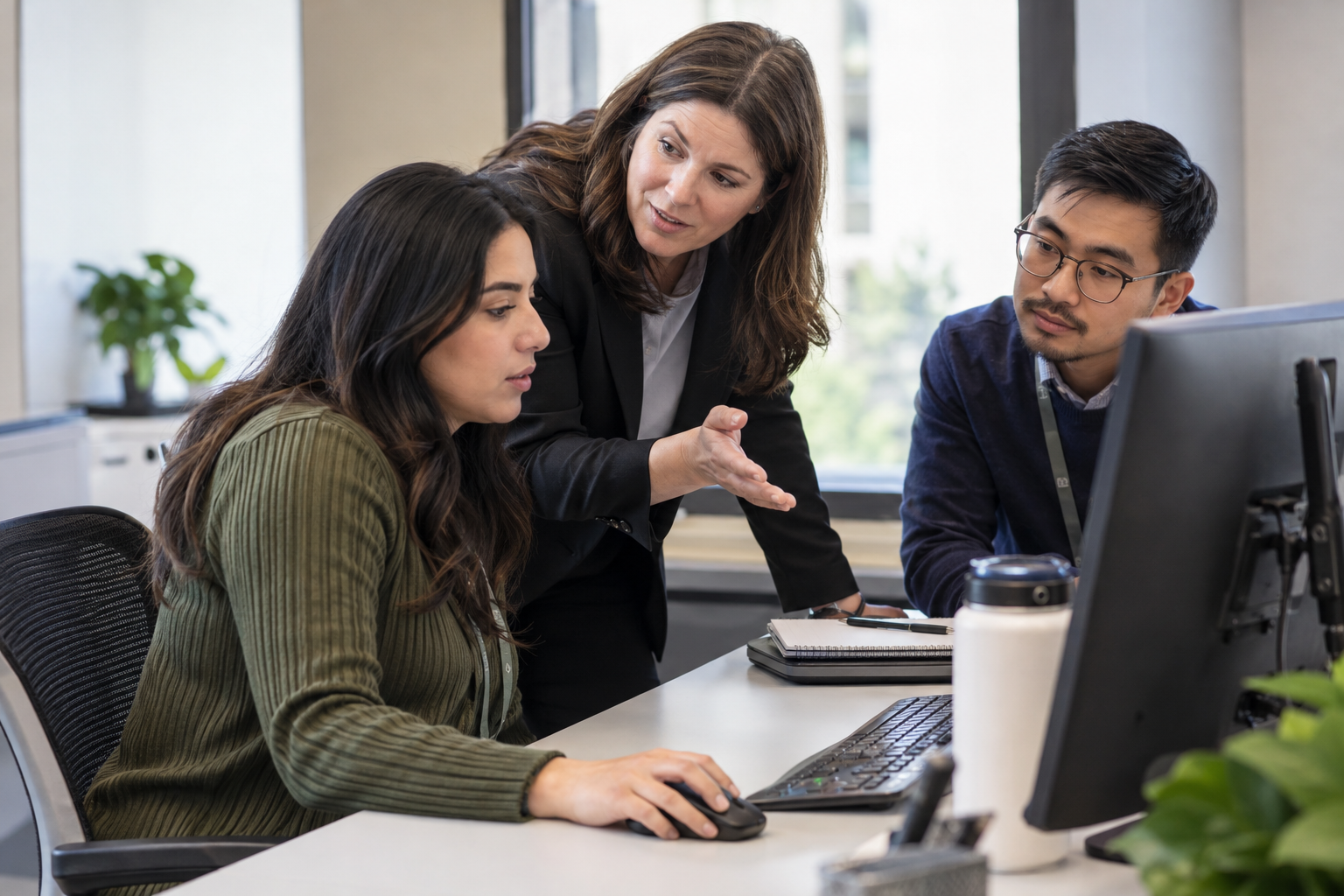 A process lead coaches an operations specialist through a human approval checkpoint at a shared workstation as a colleague listens.
