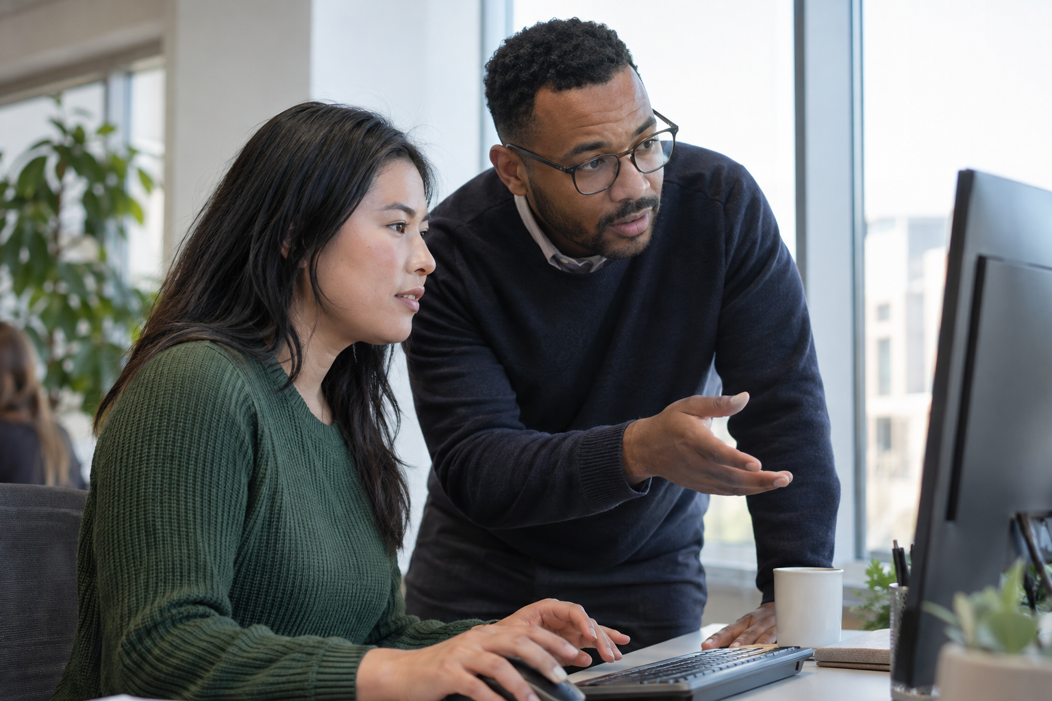 An operations supervisor and colleague discuss responsibility for an AI-assisted task while working at a desktop computer.