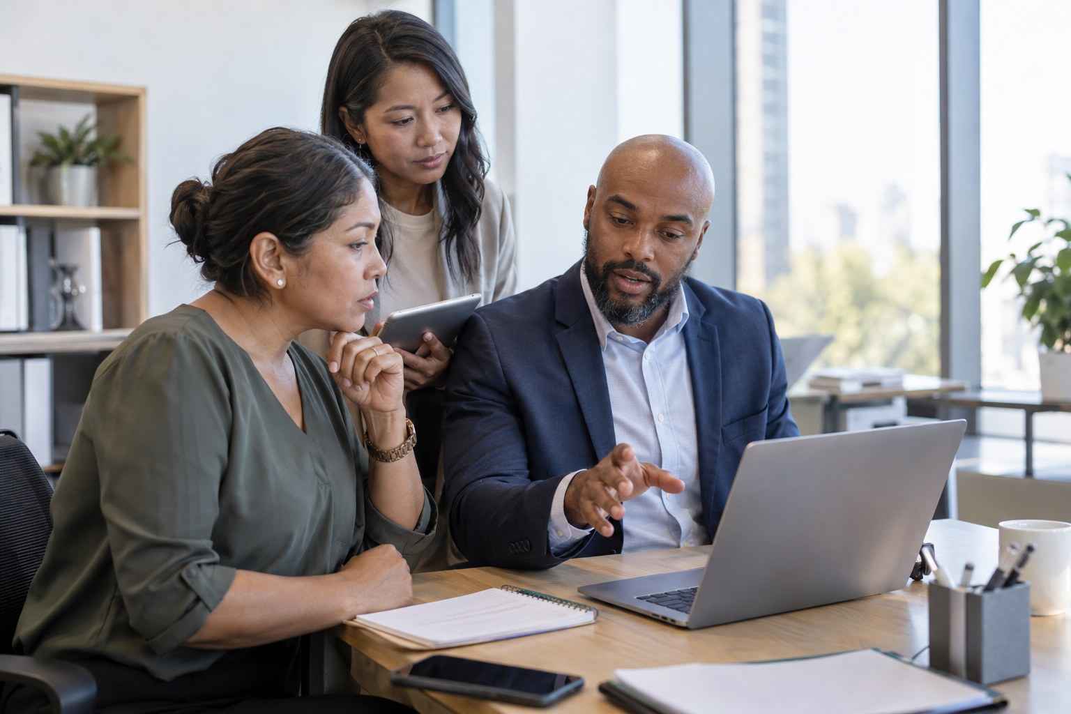 A finance manager, department lead and operations coordinator review a digital spending approval using a laptop and tablet.
