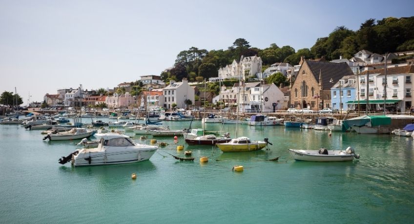 Calm green harbor with moored boats and yachts, hillside town of white and brick houses, trees, clear sky.