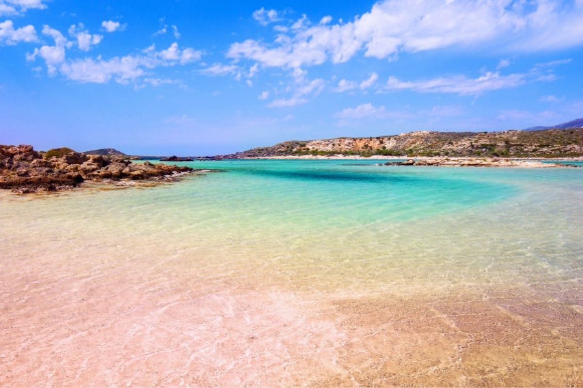 The pink grains of Elafonissi Beach in Crete and the turquoise-tinted sea and surrounding mountain landscape of Greece.