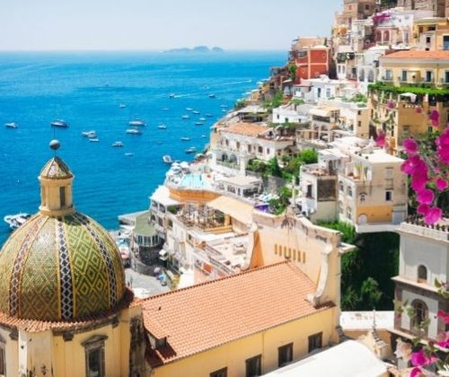 Clifftop coastal town: colorful buildings, pink bougainvillea, ornate dome, blue sea with small boats below.