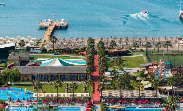 Aerial view of a beachfront resort: palm trees, pools, sun umbrellas, marina pier, boats and a speedboat on the ocean.