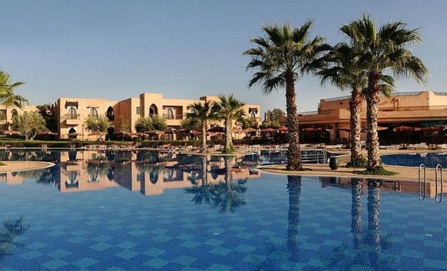 Outdoor pool with clear blue water, tall palms, resort buildings with arched doorways reflected, blue sky.