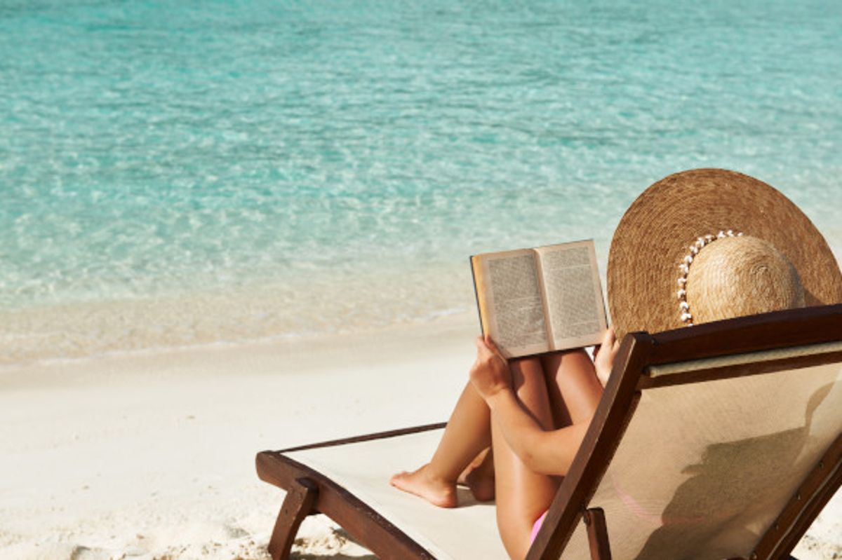Woman reading a book on a beach sunlounger with white sand and emerald waters in her view