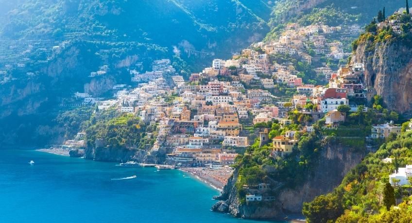 Coastal hillside town with packed colorful buildings on steep cliffs, overlooking a bright blue sea and winding shoreline.