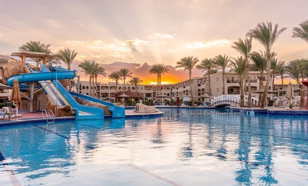 Sunset resort pool with blue water slides, palm trees, white buildings along shore, reflections on the water.