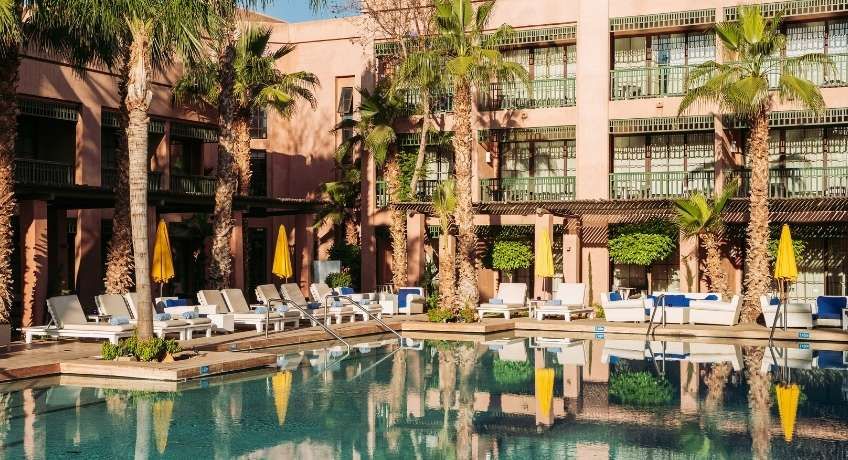 Courtyard with a reflecting pool, white lounge chairs and tables, palm trees, multi-story building with balconies.