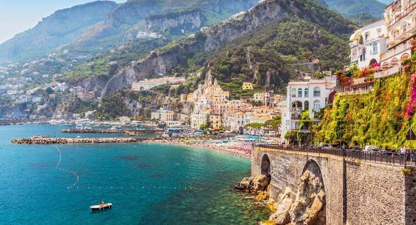 Turquoise bay with a small boat, coastal town with colorful buildings and arches, layered blue mountains, clear sky.