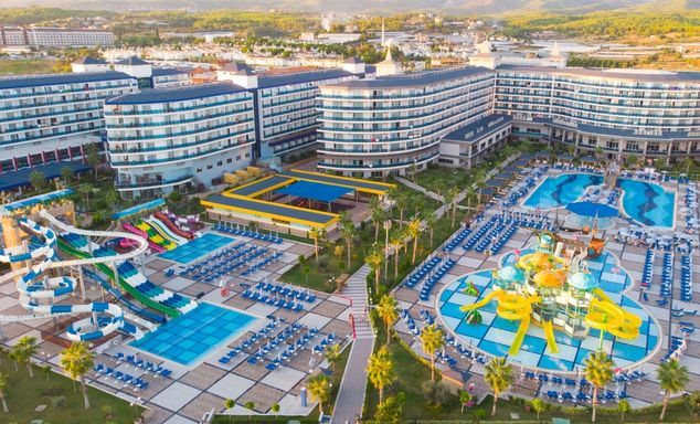 Aerial view of a large resort complex with multiple curved hotel buildings, landscaped paths, and several swimming pools including a children’s water park with colorful slides and lounge chairs.