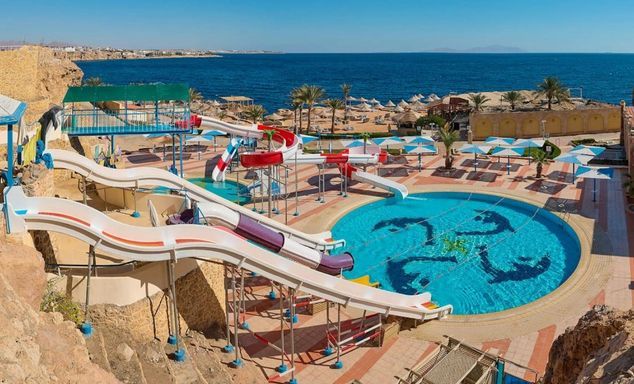 Resort water park with colourful slides above a round pool with dolphin designs, with parasols and the sea beyond.