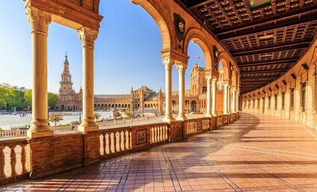 Stone colonnade with arches and carved pillars overlooks a historic skyline, tall bell tower, warm afternoon light.