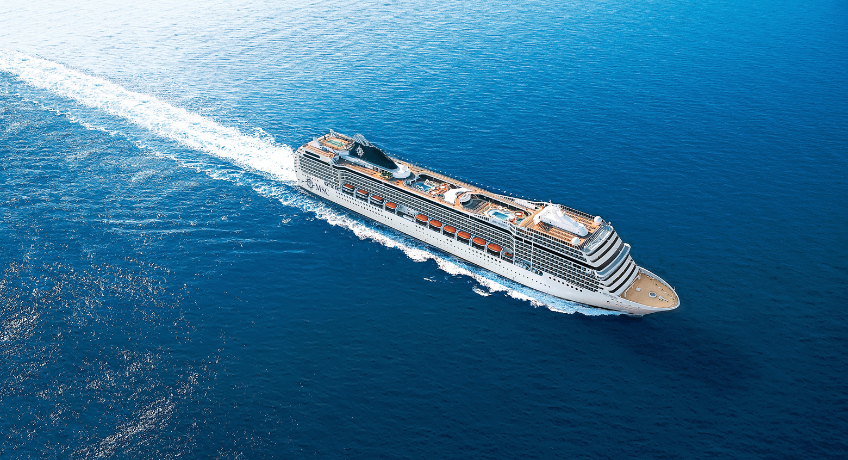 A large cruise ship sails across blue ocean water, leaving a white foamy wake behind it as seen from above.