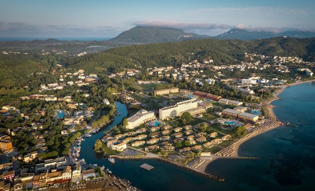Aerial view of a coastal town: curving shoreline, beaches, marinas, resort complex, green hills and mountains.