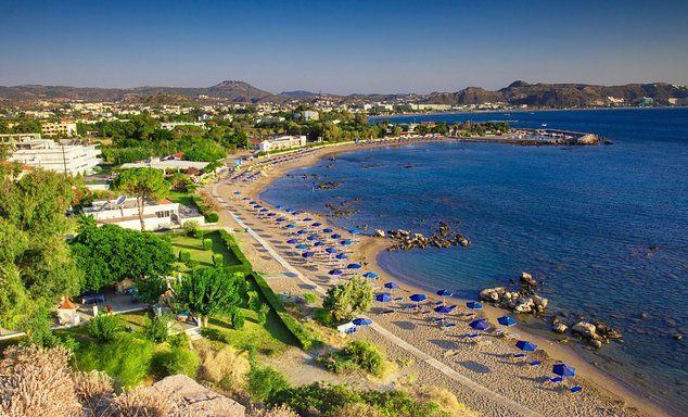 Coastal aerial view: long sandy beach with blue umbrellas, rocky shoreline, calm blue water, clear sky.