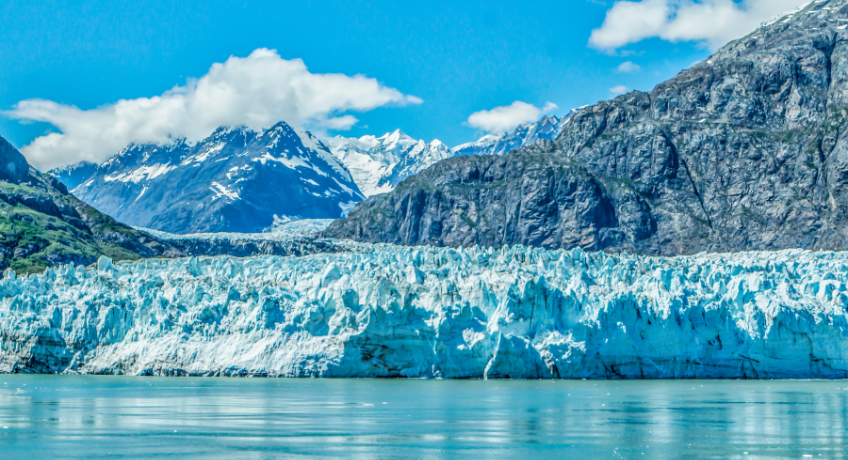 Glacial lake with calm water in the foreground, a blue ice wall and crevasses across the middle, and steep snow-capped mountains under a bright blue sky with scattered clouds.