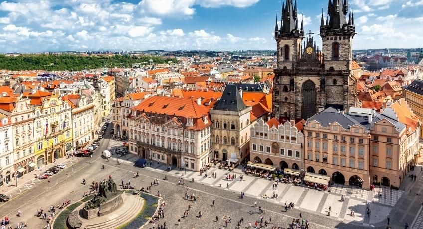 Aerial view of a historic square: terracotta rooftops, Gothic church with twin spires, people below, blue sky.