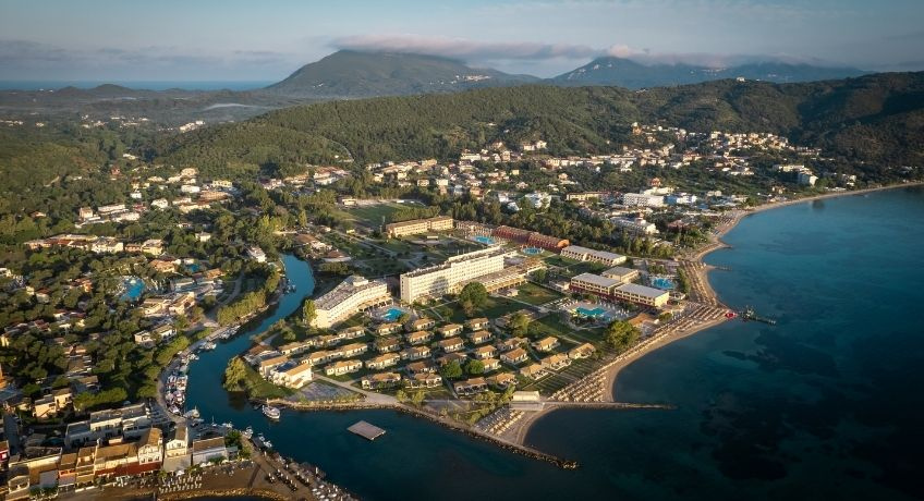High-angle aerial view of a coastal beach resort complex in Corfu, Greece, bordered by a blue bay on the right, a meandering river canal on the left, and lush green hills and mountains in the background.