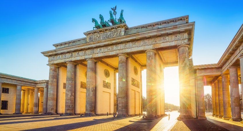 Sunrise light through columns of a grand ancient temple with a pediment and statues, cobblestone courtyard, sky.