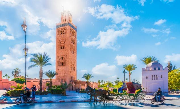 The Koutoubia Mosque's minaret rises above sandstone buildings in Marrakech, Morocco, lit by warm evening sunlight against a partly cloudy blue sky. A tree-lined pedestrian plaza with orange trees runs along the mosque's outer wall in the foreground.
