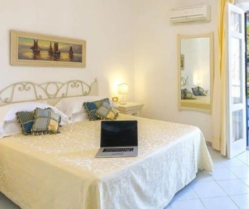 Sunlit bedroom: made bed, patterned pillows, bedside lamp, open laptop, yellow-curtained door to a terrace with seating.