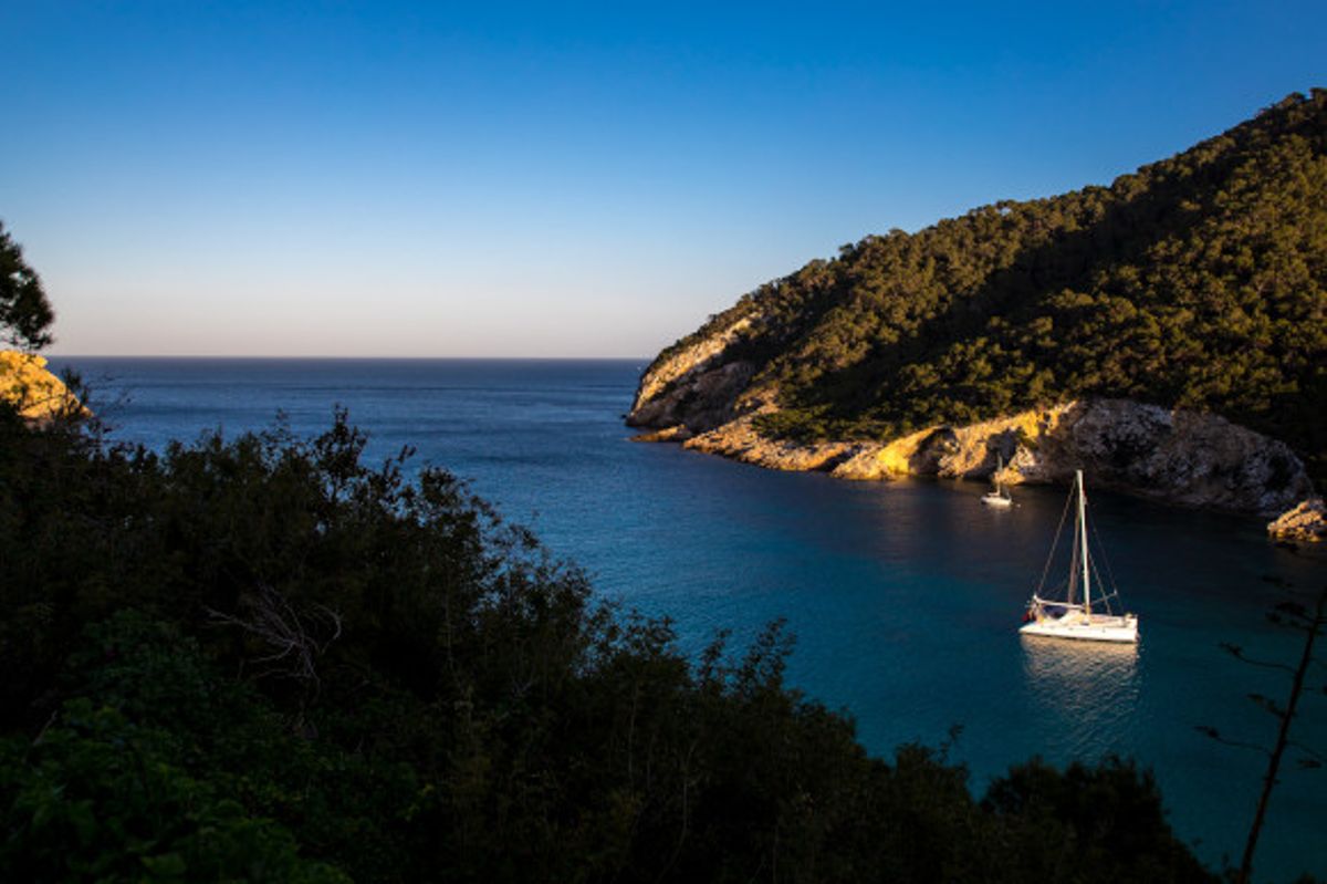A picturesque beach in Cala Llonga, Ibiza with a boat in the water and green cliffs lit up by the sun