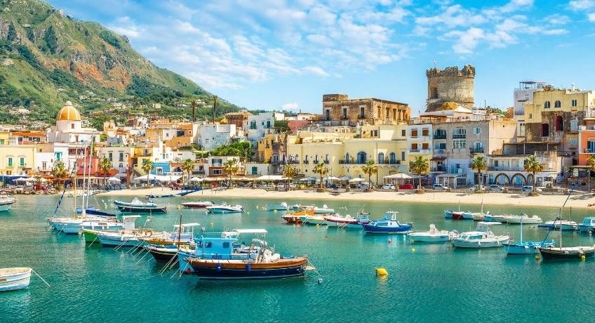 Harbor of moored boats and yachts, coastal town with colorful buildings, stone church tower, green hills, blue sky.
