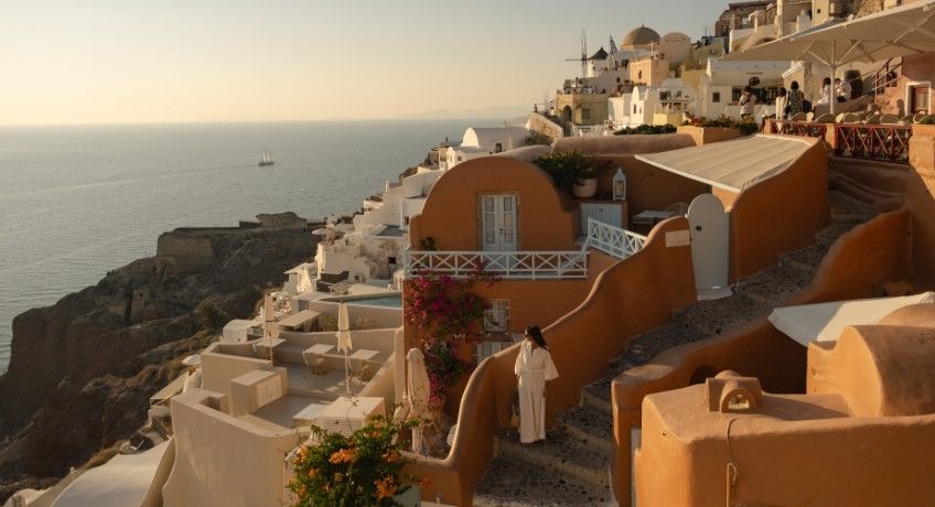 Terraced white buildings with orange domes and balconies overlook a calm sea at sunset, with a small boat on the water and a few people walking along the steps.