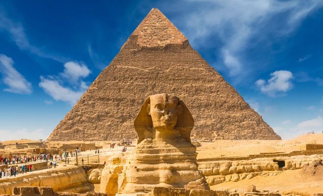 Great Pyramid of Giza with the Sphinx in front, sandy ruins, visitors along the walkway, blue sky with clouds.