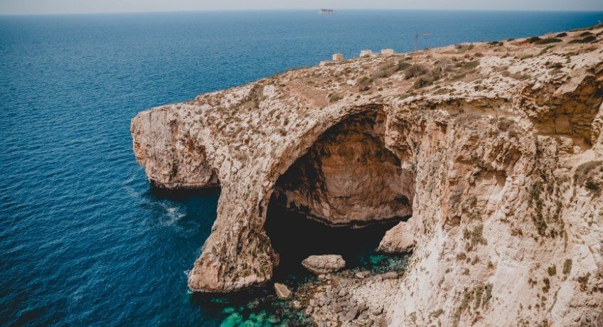 Cliffside limestone arch over turquoise waves, rugged beige rock, distant ship on the horizon, hazy blue sky.