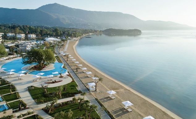 Aerial view of a beachfront resort with pools, gardens and white parasols along a sandy shore, with calm sea and mountains.