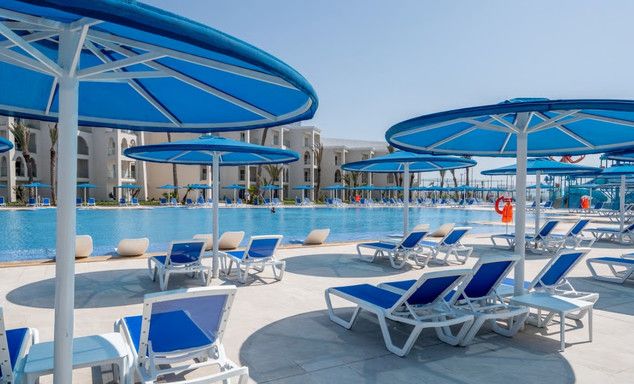 Poolside terrace: rows of blue-white lounge chairs and umbrellas, large pool, low-rise buildings, clear sky.