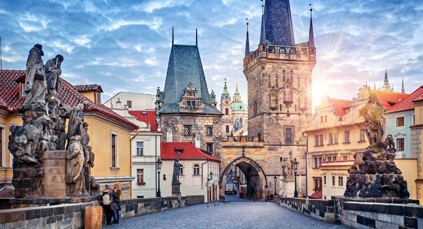 Historic European street: cobblestones, red-tiled buildings, stone archway gate, Gothic towers and spires, sky.