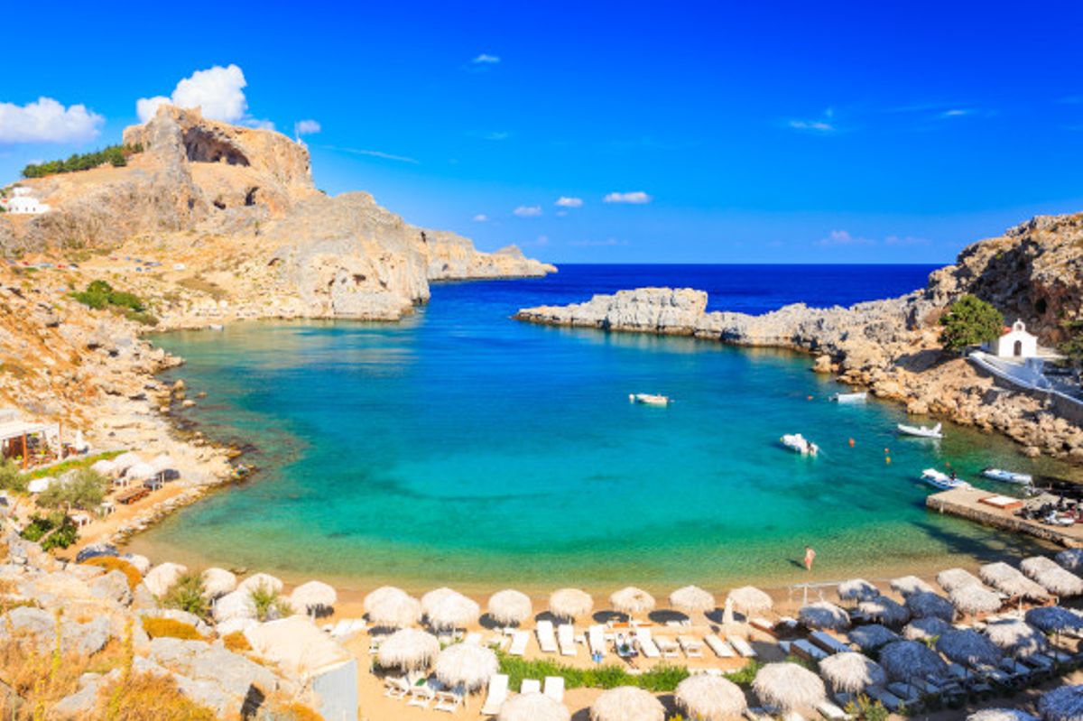 St. Paul’s Bay in Rhodes lined with sunloungers with a traditional chapel on the right hand side of the shore