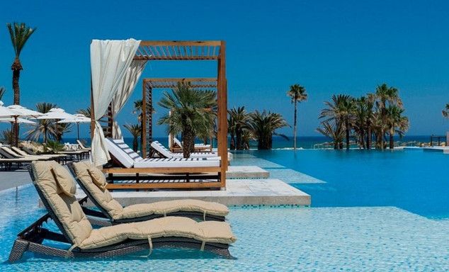 Beige cushioned loungers beside a blue mosaic pool, wooden pergolas, palm trees, calm ocean and blue sky.