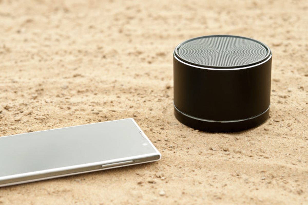 Black Bluetooth speaker on the sand with sliver mobile phone next to it