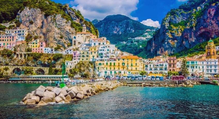 Coastal town with colorful buildings on a rocky shoreline, breakwater stones, turquoise water, green mountains, cloudy sky.