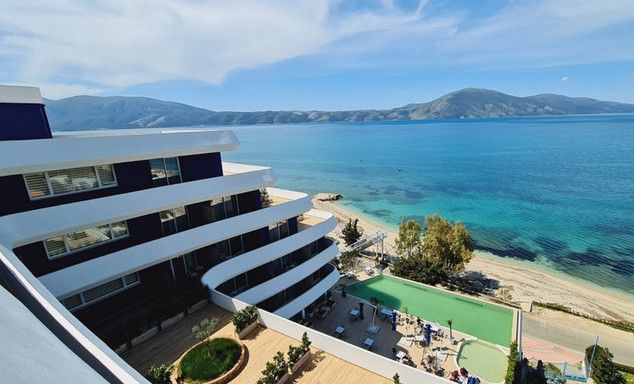 Aerial view of a beachfront resort: balconies, turquoise ocean, distant hills, and a small pool with sunbeds on the sand.