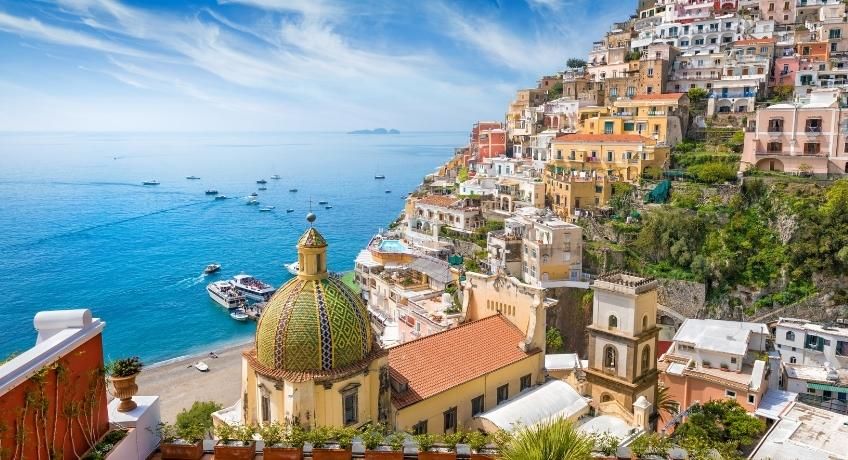 Hillside coastal town with colorful buildings, tiled church roof, golden dome terrace, blue sea, boats, distant horizon.