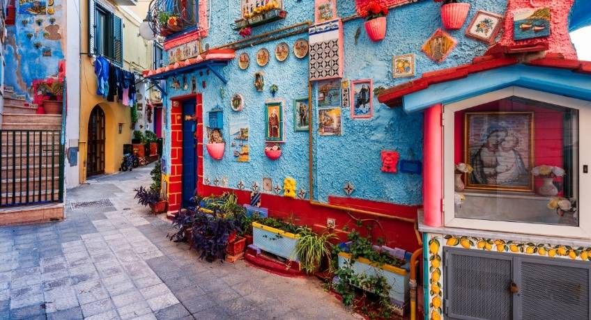 Narrow street with blue walls, murals, framed pictures, plants, hanging red lanterns, and storefronts in daylight.