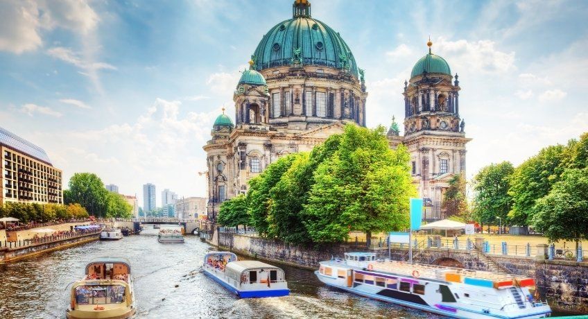 River view toward a grand domed cathedral with two towers, green riverbanks, sightseeing boats, blue sky.