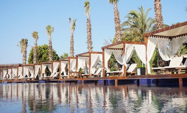 Lakeside event deck: wooden chairs, white draped canopy, reflected in still water, palm trees, clear blue sky.