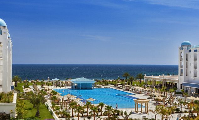 A large outdoor swimming pool surrounded by palm trees leading to blue sea water