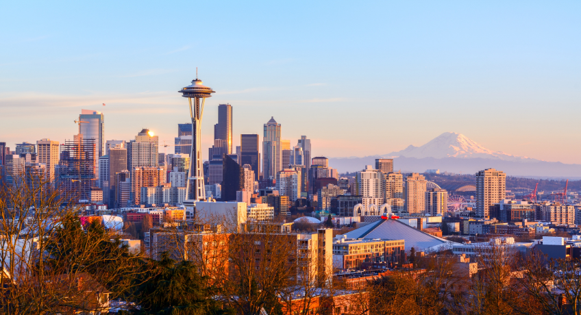 Seattle skyline at dusk with the Space Needle, tall downtown buildings, and distant snow-capped Mount Rainier under a pastel blue sky.