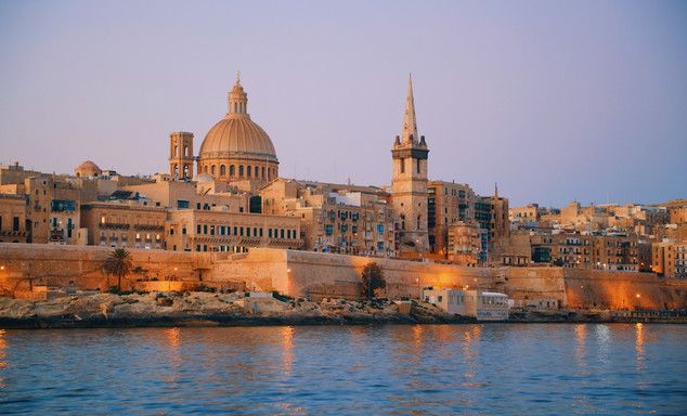 Dusk view of a historic waterfront city: domed cathedral, tall bell tower, clustered stone buildings, calm water.
