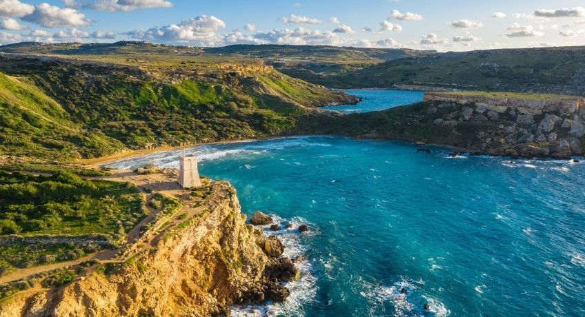 Aerial view of a turquoise coastline: rocky cliffs, winding coastal road, small waves, blue sky with clouds.