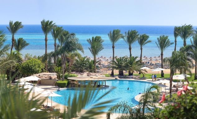 Resort pool with a swim-up bar surrounded by palm trees, with a sandy beach lined with umbrellas and turquoise sea beyond.